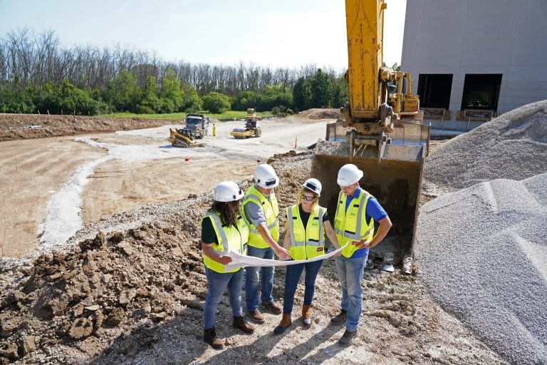 Construction team wearing safety vests and hard hats reviewing blueprints at an active job site with heavy equipment and earthwork in progress.
